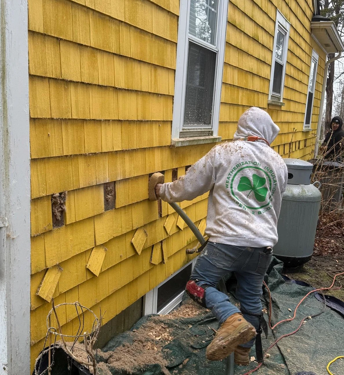 McMahon Insulation crew dense-packing a wall cavity from outside the house, with a course of shingles removed for access